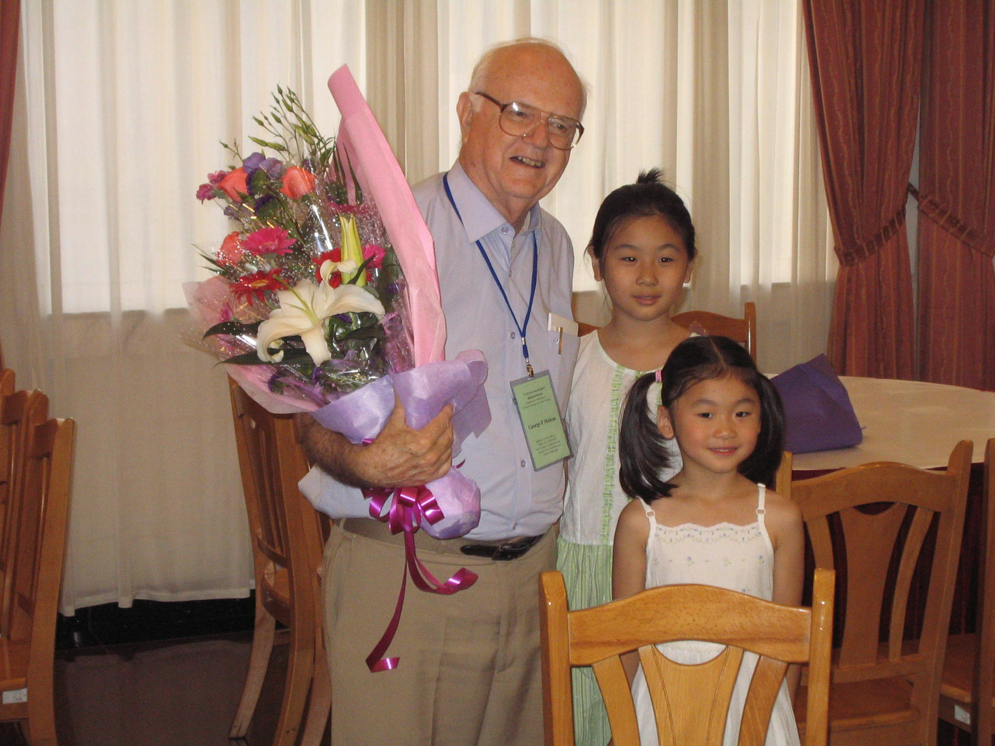 Father McLean smiling warmly holding a colorful floral bouquet presented by children at his 75th birthday